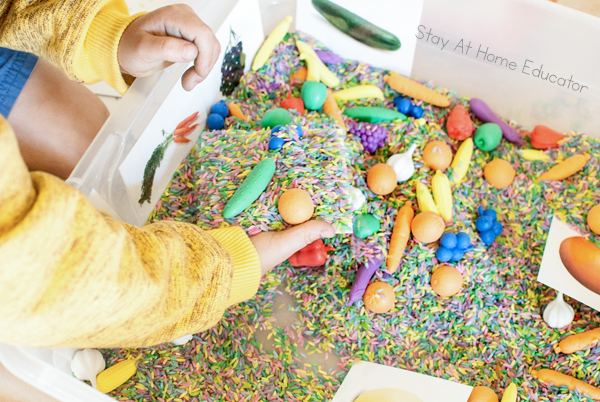 Image shows preschoolers engaging with a fruit and veggie sensory bin for preschoolers| The image includes a clear bin which contains colored rice, fruit and veggie mini manipulatives, and fruit and veggie cards from a free printable|