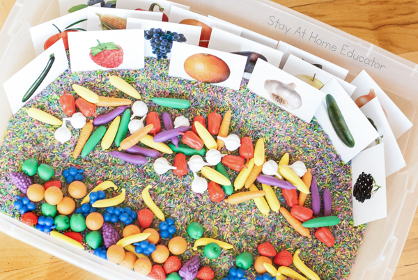 Image shows fruit and veggie sensory bin for preschoolers| The sensory bin image includes a clear bin holding colored rice, fruit and veggie mini manipulatives, and fruit and veggie cards from a free printable|