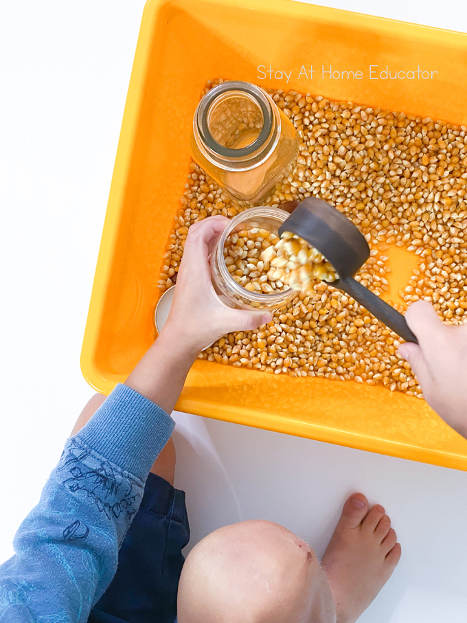 scooping and pouring activity for toddlers| sensory bin with corn kernels| child's hand is pouring corn kernels from a glass jar to a small clear container| farm theme scooping activity for toddlers|
