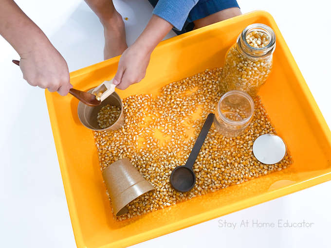 scooping and pouring activity for toddlers| corn kernel sensory bin| a variety of small containers and scoops| a child is using two different scoops for scooping and pouring|
