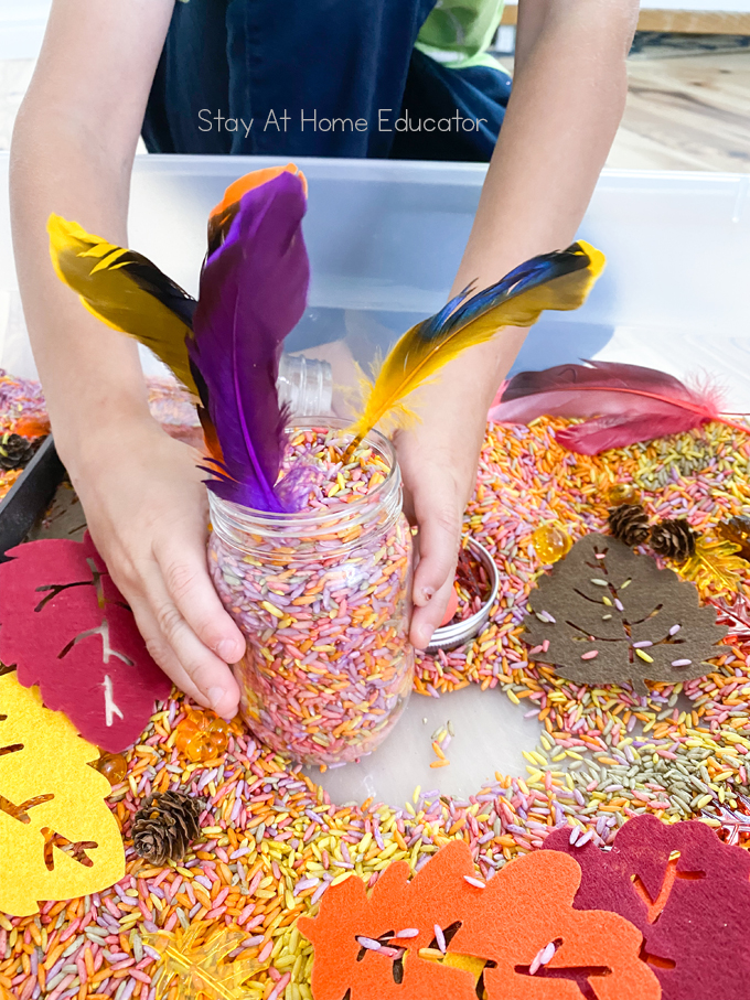hands holding a jar filled with colorful rice and feathers stuck in it like a vase inside a fall themed sensory bin