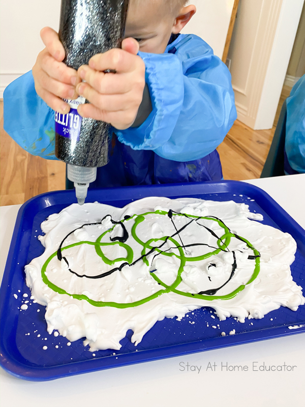 a preschooler squeezing glitter glue all over his shaving cream tray as he finishes a fun winter art project for preschoolers