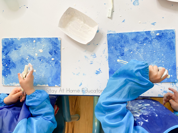 two children using a toothbrush to splatter white paint on their winter landscape art activity