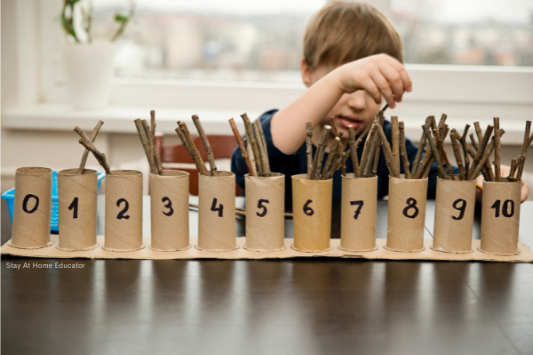 toilet paper tubes with numbers written on and the corresponding number of sticks inside and a boy practicing one to one correspondence