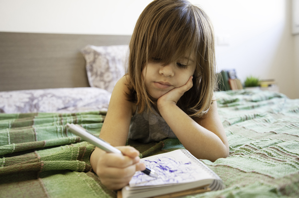 A young girl scribbling in a notebook on her bed is an important step of emergent writing activities for preschoolers