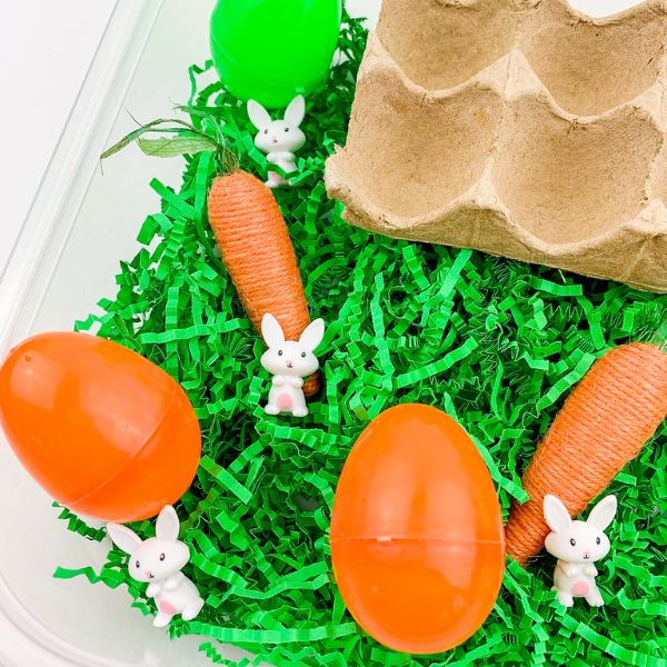 green grass, orange carrots, and white bunnies mix with colorful easter eggs in an easter grass sensory bin