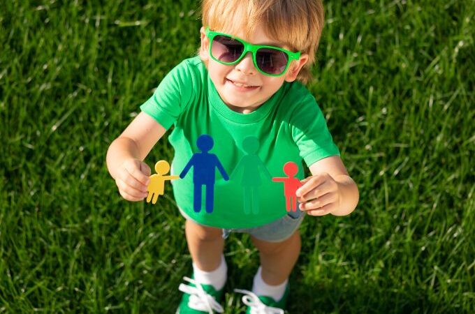 Young boy outside holding up paper cutouts of kids smiling at the camera | All About me preschool lesson plans | September preschool themes |