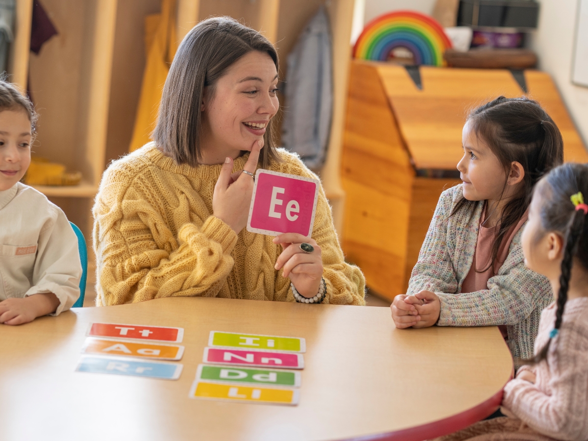 letter recognition objectives include: visual discrimination, letter naming, letter formation, and letter sound correspondence | teacher sitting with children, holing up a letter E flashcard, and pointing to her mouth as she says the letter