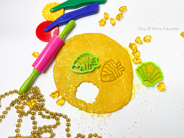 super soft playdough recipe using gold loose powder eye makeup and chunky gold glitter | playdough for St. Patrick's Day or pirates preschool theme | playdough pancake with rolling pin, playdough cutters, fish and sea shell cookie cutters and gold necklace