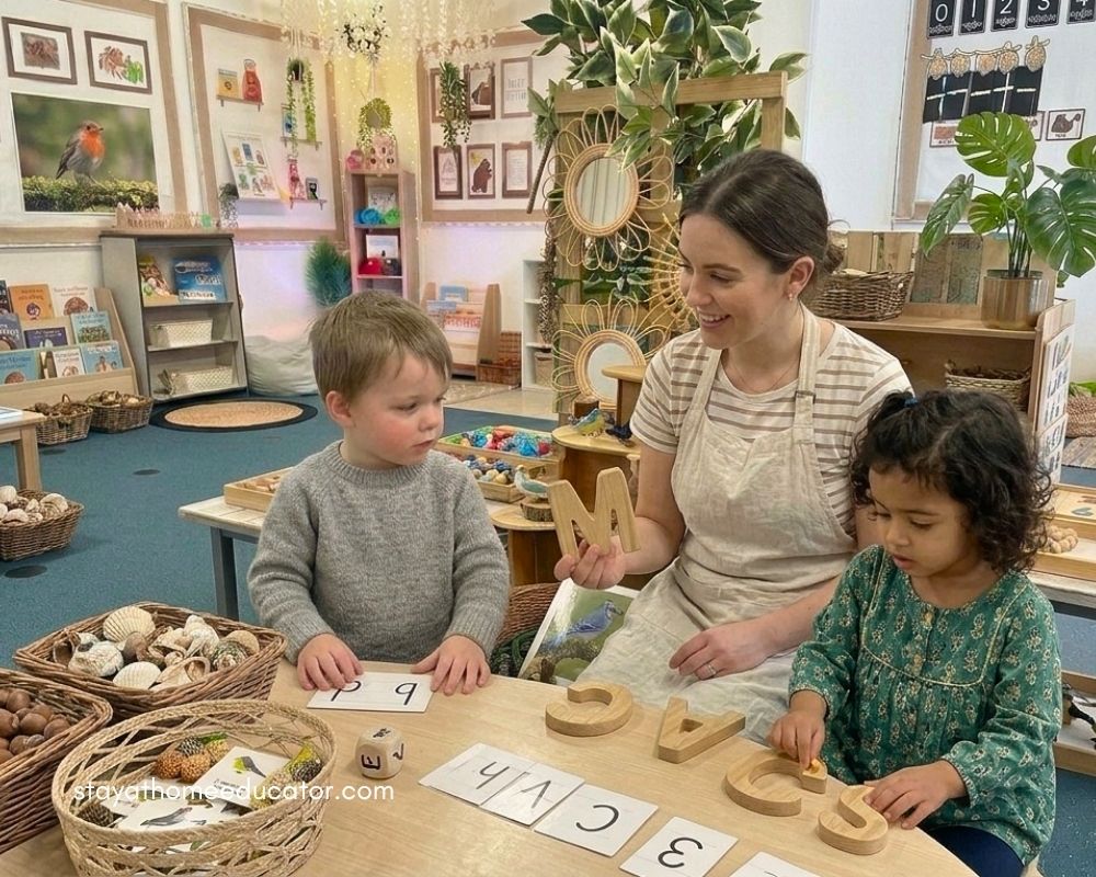 Preschool teacher supports two children using wooden letters and cards to practice letter recognition and phonics skills through hands on, play based learning in a natural classroom environment.